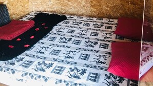 Una habitación pequeña y acogedora con techo y paredes de madera, cubiertas de virutas. Hay una cama con una colcha estampada y dos almohadas rojas.