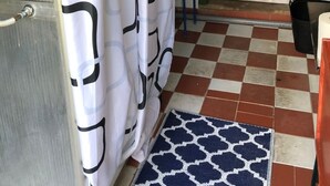 Un baño con piso a cuadros, cortina de ducha con diseño geométrico, una puerta verde y una silla azul.