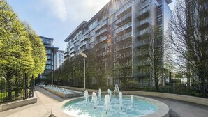 A modern urban setting with a central fountain, surrounded by trees and a multi-story building with balconies. - Luxury Apartment at Chelsea Bridge/Battersea Power Station (London)
