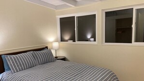Un dormitorio con cama, mesita de noche, lámpara y dos ventanas.