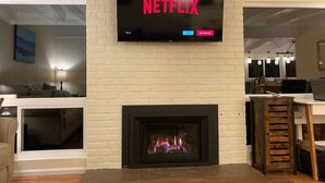 Un salón con chimenea, un televisor montado encima mostrando el logotipo de Netflix, y un ordenador portátil sobre una mesa a la derecha.