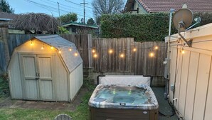Un patio trasero con un jacuzzi, dos sillas Adirondack, un cobertizo y una guirnalda de luces.