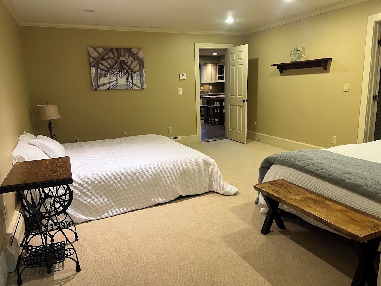 Une chambre avec deux lits, une machine à coudre, un banc, un tableau et un coin cuisine visible à travers une porte ouverte.