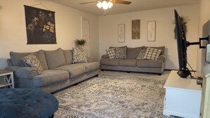 Un salon avec deux canapés gris, des coussins à motifs, un grand tableau représentant des fleurs, un élément décoratif mural et un téléviseur à écran plat.