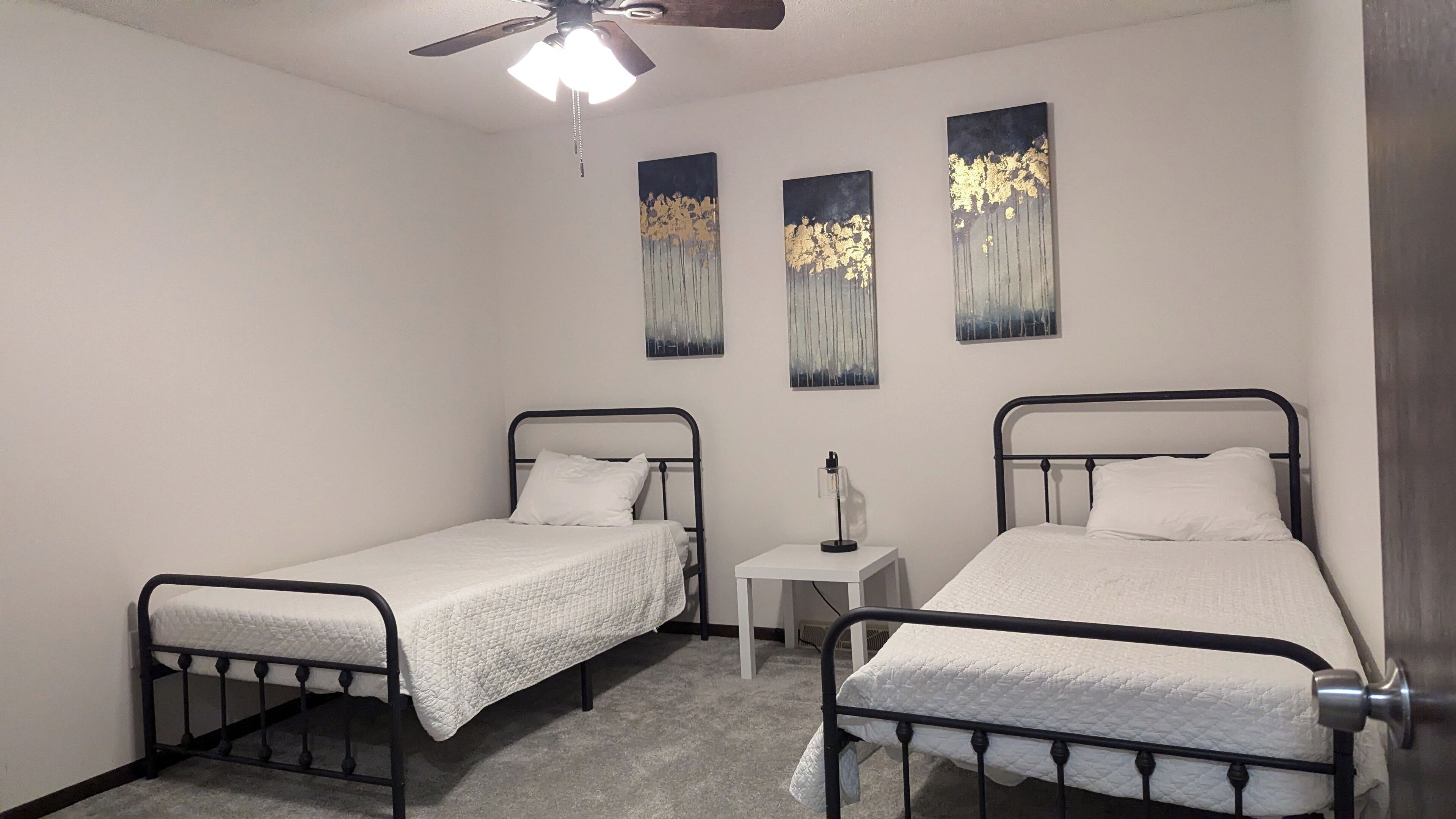 Un dormitorio con dos camas individuales, un ventilador de techo y tres cuadros colgados en la pared.