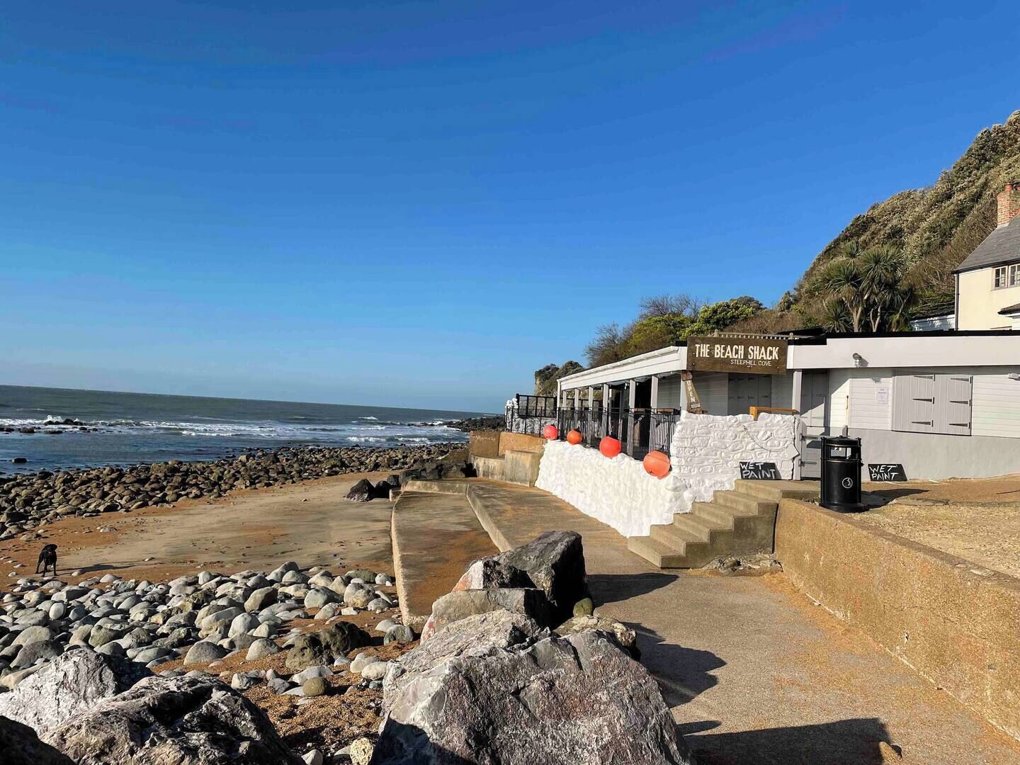 A coastal building named "The Beach Shack" with a stone facade, steps leading to a covered entrance, and a view of the ocean.