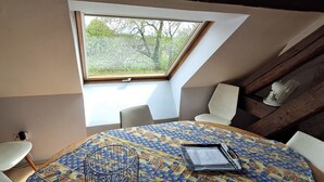 Une petite pièce agréable, dotée d’un plafond en pente et d’un puits de lumière, comprenant une table ronde avec des chaises, un présentoir métallique et un petit bureau avec des papiers.