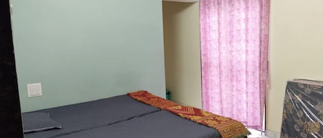 A single bed with a grey mattress and a patterned blanket, a power outlet on the wall, and a pink curtain on the window.