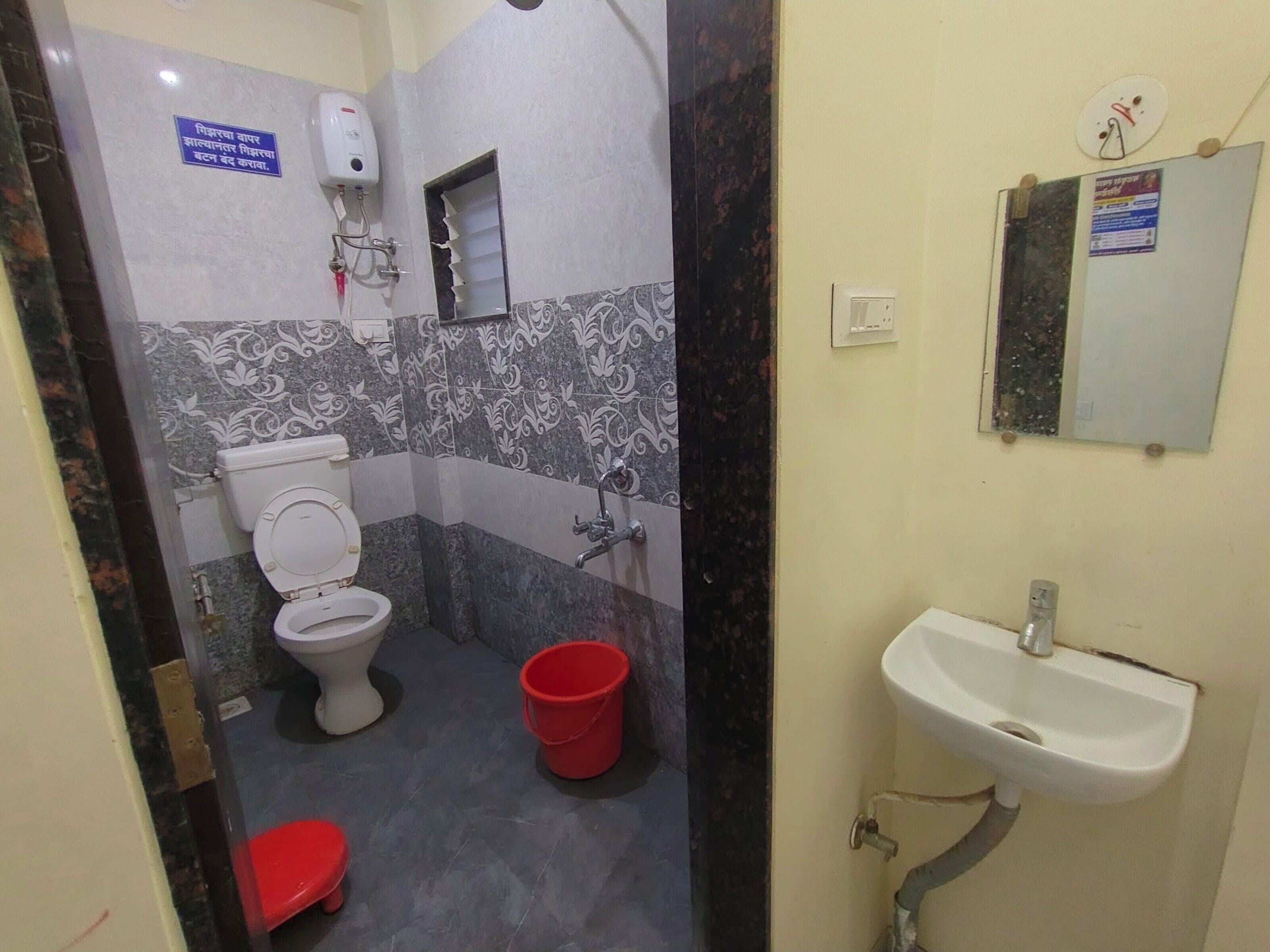 Ein Badezimmer mit Toilette, einem roten Eimer, einem Waschbecken und einem Spiegel.