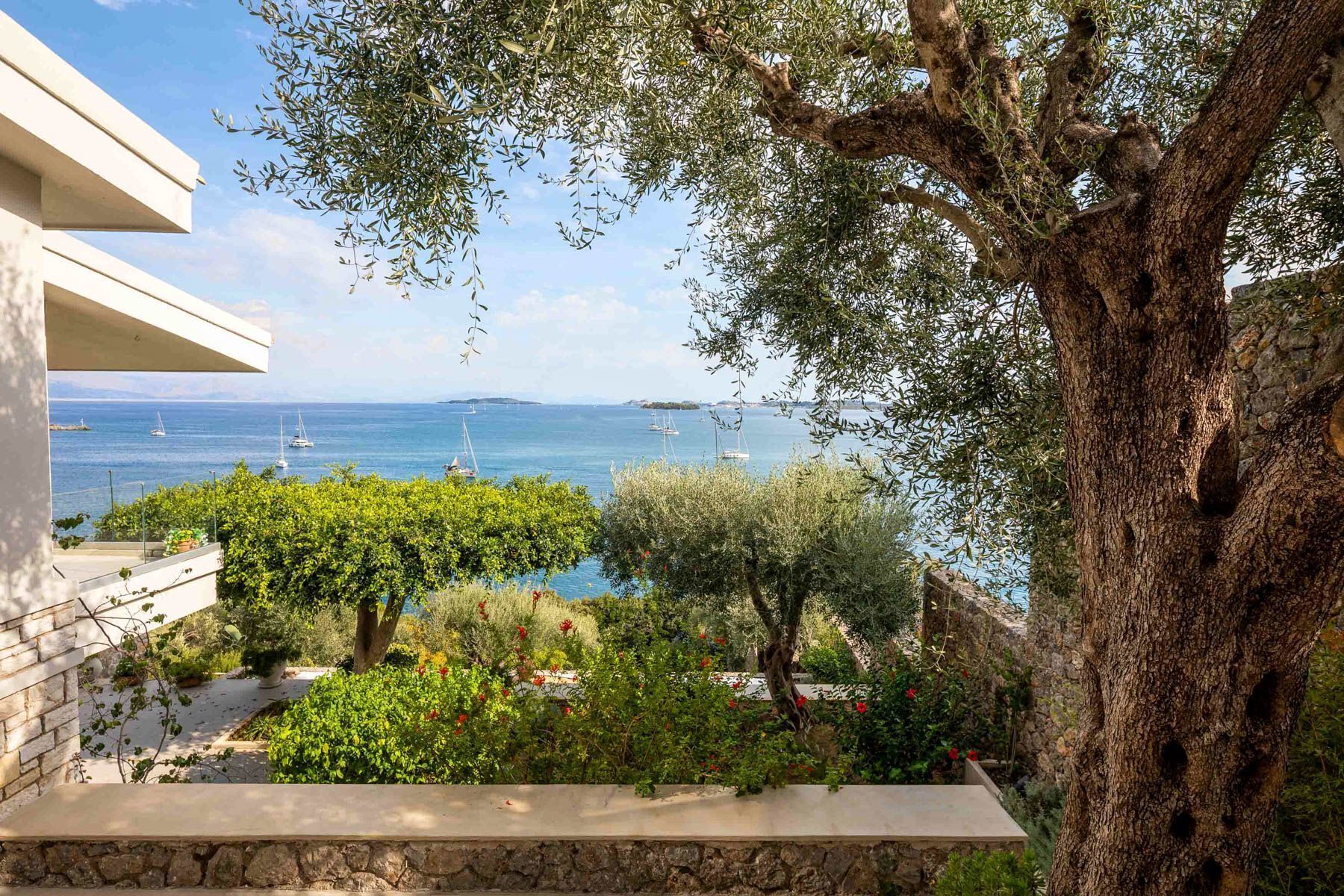 A balcony with a stone wall, olive trees, and a view of the sea with sailboats.