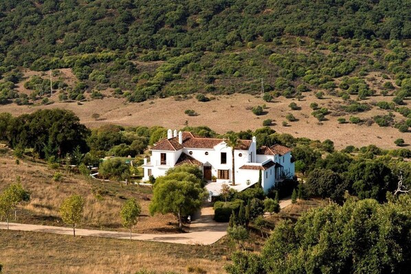 A white villa with a tiled roof, surrounded by greenery and trees, set on a hillside.