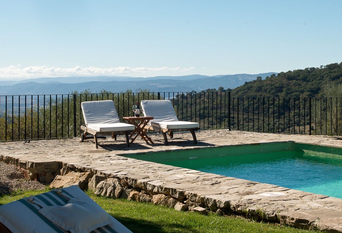 A pool area with two lounge chairs and a table, surrounded by stone walls and a metal fence, overlooking a mountainous landscape.