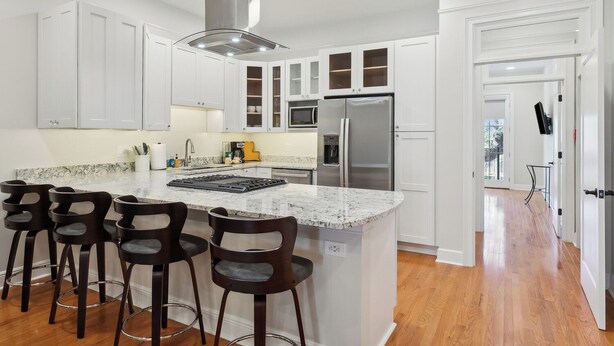A modern kitchen with white cabinets, a central island with bar stools, stainless steel appliances, and a view to the outside.