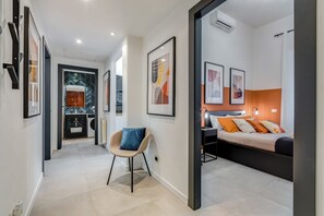 Een moderne hotelkamer met een bed, nachtkastje en een stoel. Er is een gang die leidt naar een badkamer met een spiegel en wastafel.