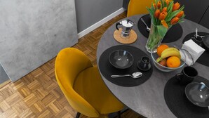 Een moderne eetruimte met een ronde tafel, zwarte en gele stoelen, een koffiepot, een fruitschaal en een vaas tulpen.