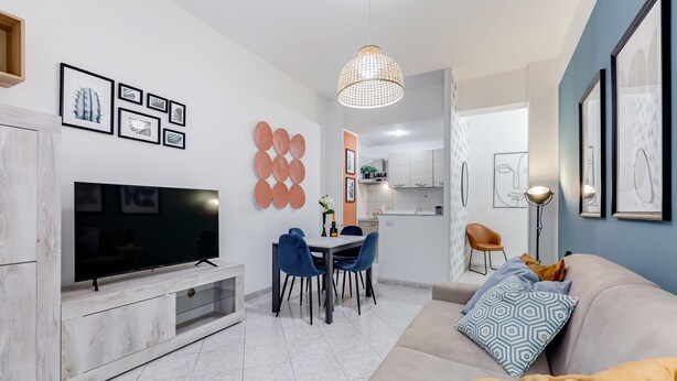 Een moderne woonkamer met een flatscreen televisie, een eethoek en een bank met decoratieve kussens.
