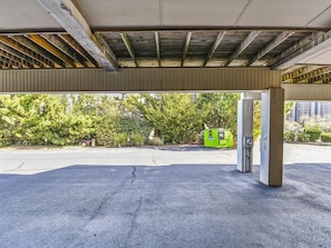 An empty parking area with a concrete structure overhead, a green trash bin, and trees in the background.