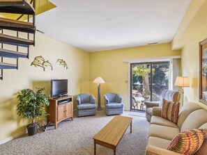 Una sala de estar con un sofá, dos sillones, una mesa de centro, un televisor sobre un mueble de madera, una lámpara y una escalera.