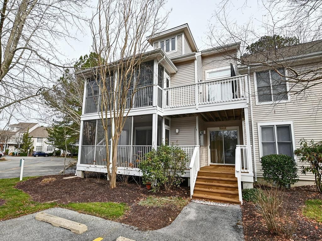 A two-story residential building with a balcony on the upper floor, a front porch, and a small front yard with a tree and shrubs.