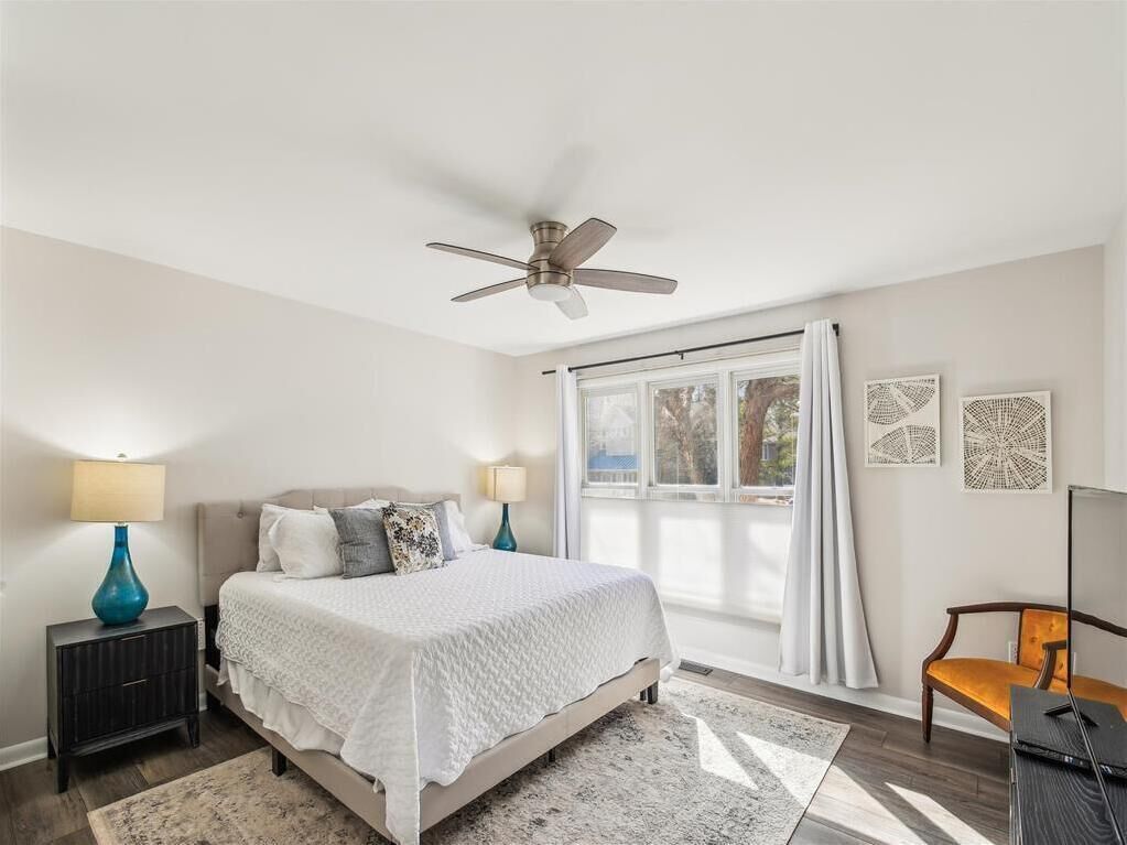 Une chambre à coucher comprenant un lit, des tables de chevet, un ventilateur de plafond, une fenêtre avec des rideaux et des œuvres d’art murales.