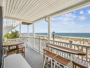 A covered outdoor deck with wooden tables and chairs, overlooking a beach with a clear view of the ocean. - Oceanfront 1BR w/ Balcony & Sea Views in Bethany (Bethany Beach)