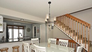 A dining area with a glass table and white chairs, a kitchen with a window, and a wooden staircase.