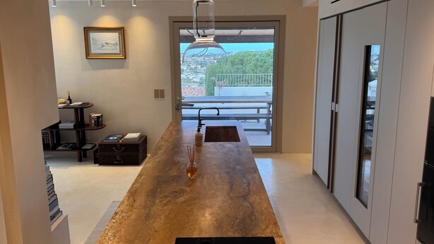 A modern kitchen with a central island, a dining area with a bench, and a view of a cityscape through a large window. - Eclat de Provence - luxury villa - calanques de cassis - (Allauch)