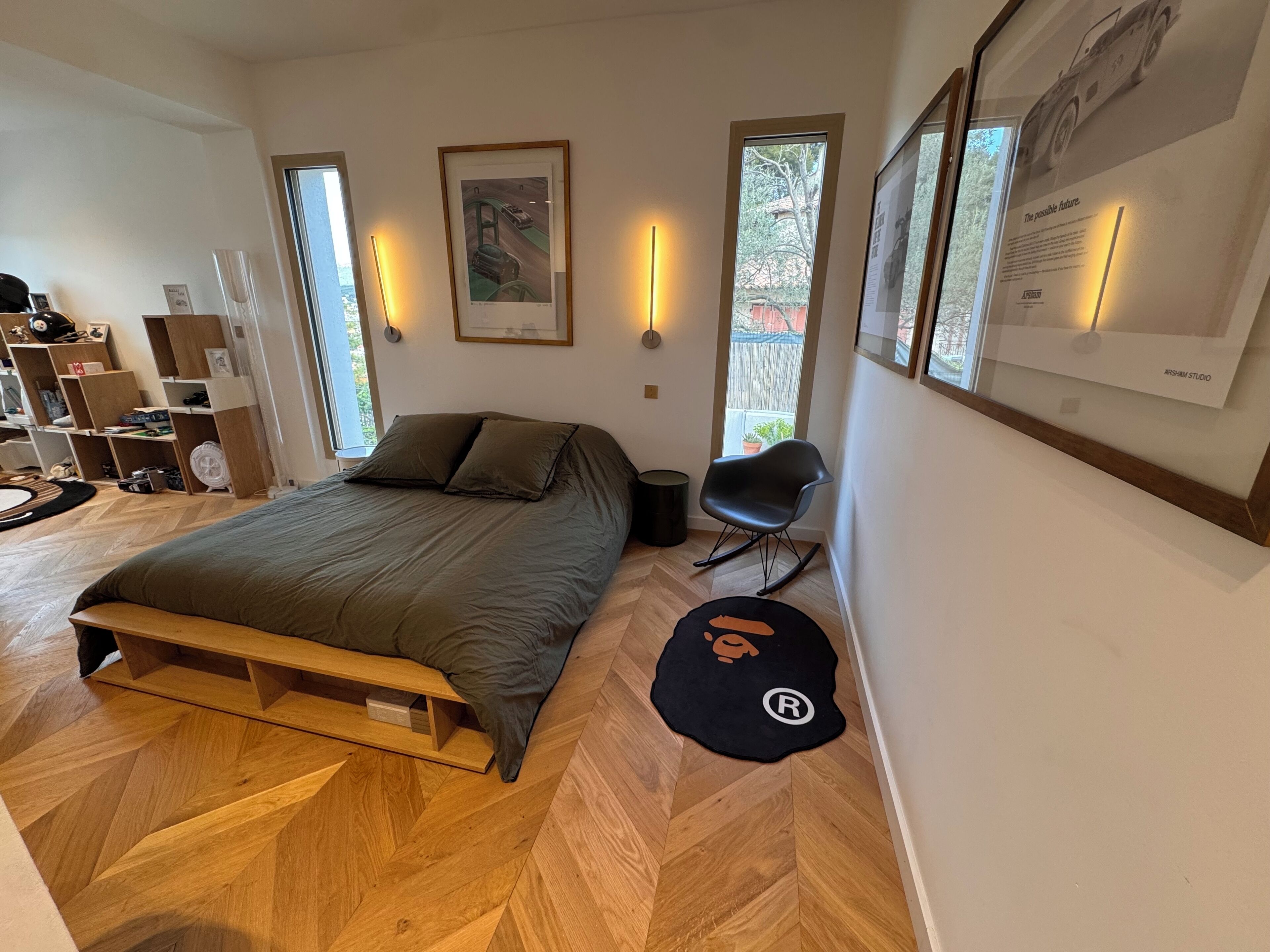 A modern bedroom with a wooden floor, a bed with a wooden frame, a rocking chair, and a black rug with a logo.