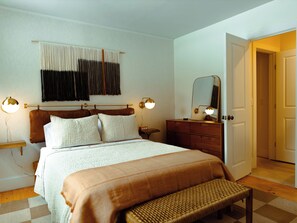 Um quarto com cama, mesas de cabeceira, penteadeira, um banco, um espelho e uma porta para outro cômodo.