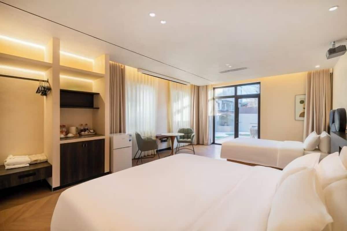 Quarto de hotel com duas camas, uma escrivaninha, uma cadeira, uma mesinha e uma janela com cortinas.