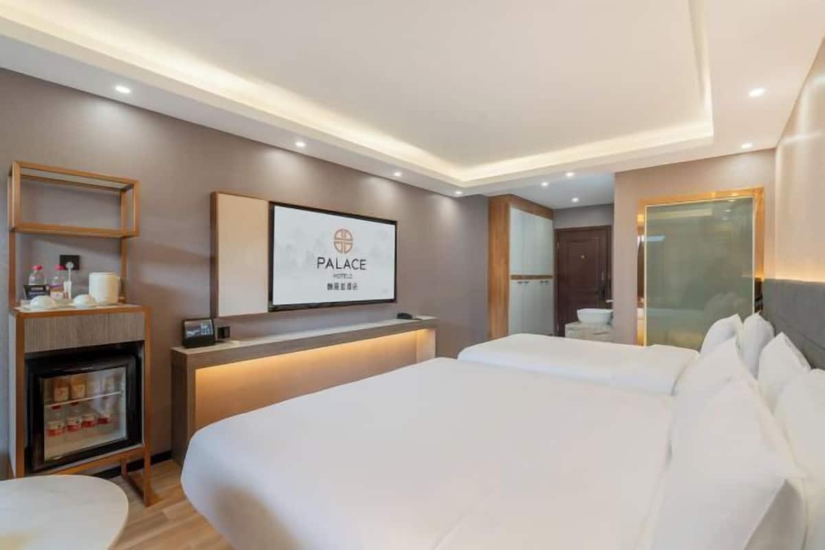 Quarto de hotel com cama grande, mesa de cabeceira, pequena geladeira, espelho e televisão presa à parede.