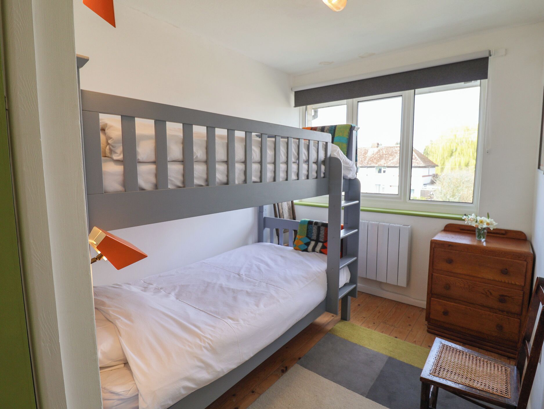 A bunk bed with a lower bunk, a wooden dresser, a chair, and a window with a view of greenery.