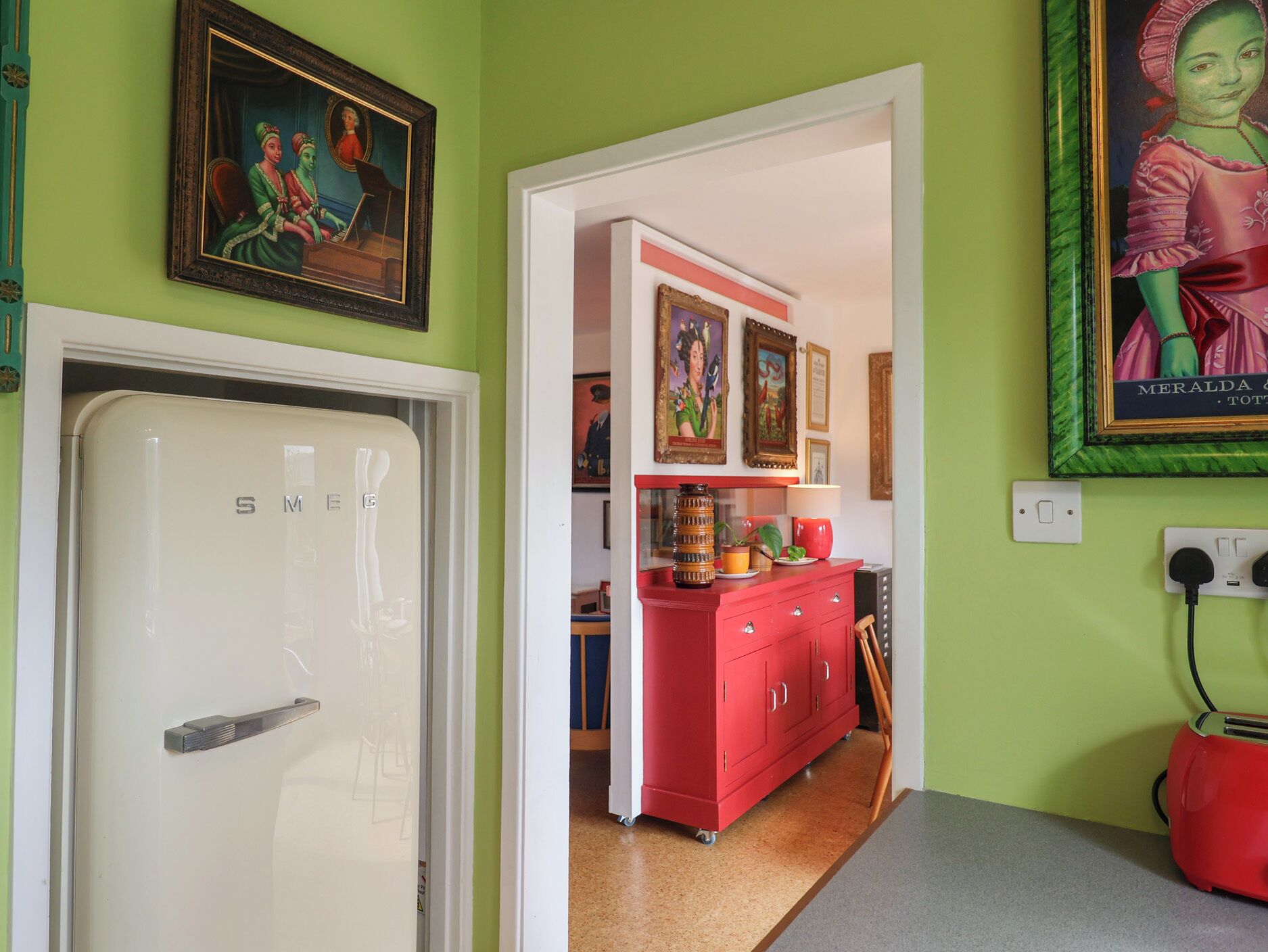 A kitchen with a white refrigerator, a red cabinet, and various framed artworks on the walls.