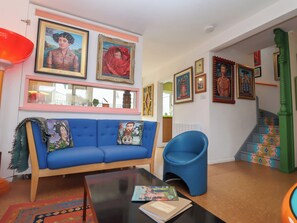 A living room with a blue sofa, a blue armchair, and a coffee table with books. There are framed pictures on the wall and a staircase with colorful steps. - The Orchard Close Art House (Bury St Edmunds)