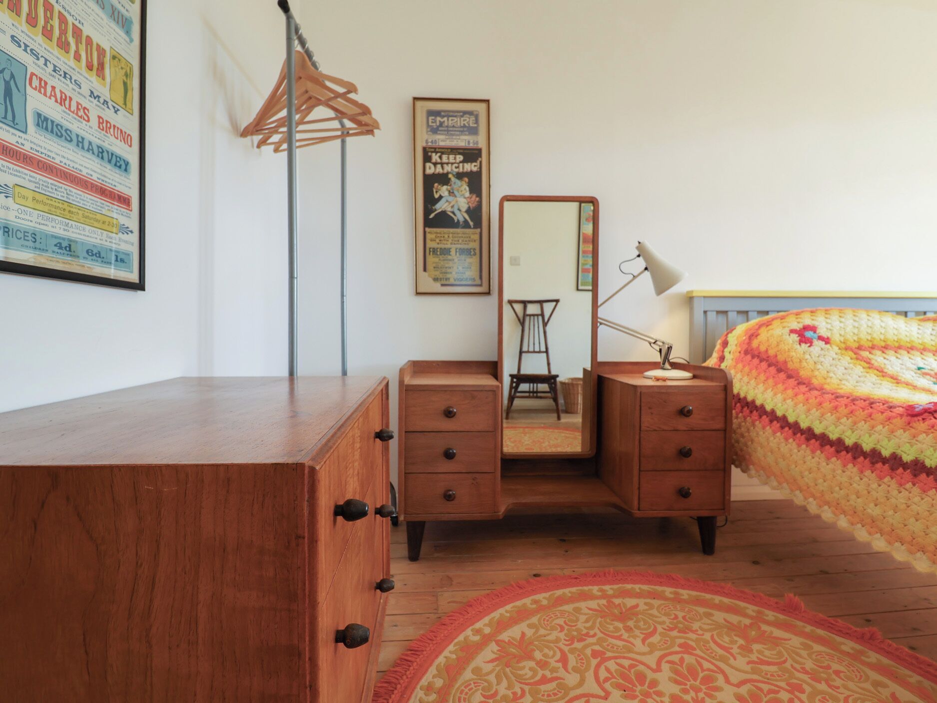 A bedroom with a wooden dresser, a mirror, a desk with a lamp, and a colorful blanket.