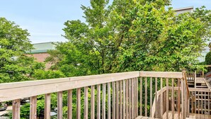 A wooden deck with a railing, surrounded by lush green trees and a building in the background.
