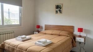 Ein ordentlich gemachtes Bett mit einer braunen Tagesdecke und zwei weißen Handtüchern, ein hölzernes Kopfteil, zwei Nachttische mit roten Lampen, eine Heizung und ein Fenster mit Blick auf Grünflächen.