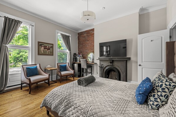 A bedroom with a large bed, a desk, a chair, a fireplace, and a TV. - Blue George Suite Historic DT (Halifax)