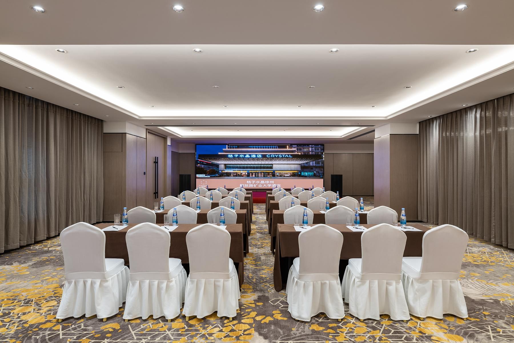 A conference room with rows of tables and chairs, a large screen at the front, and a patterned carpet.