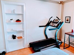 A room with a treadmill, a white shelving unit with various items, and a small table with a picture frame.