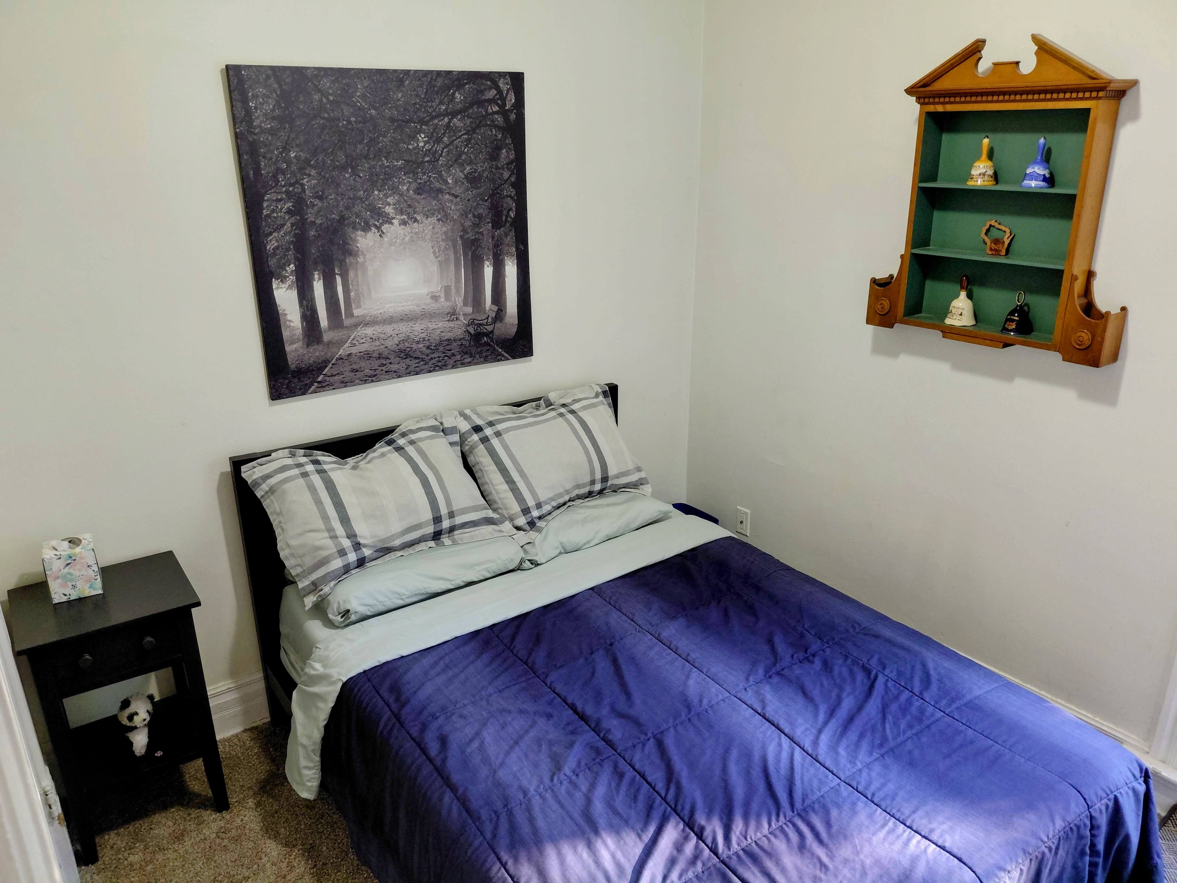 A bedroom with a bed, a nightstand, a wall-mounted shelf with decorative items, and a framed picture of a forest path.