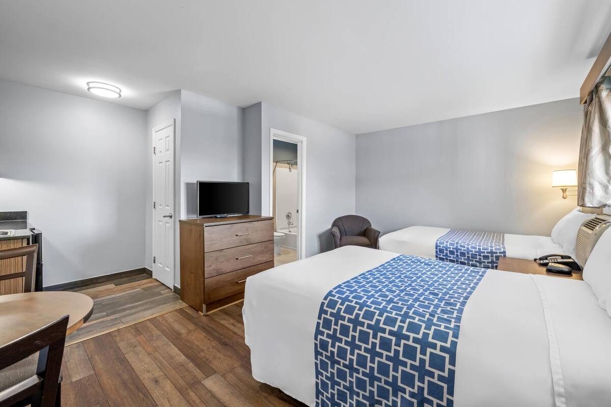 Quarto de hotel com duas camas, uma escrivaninha, uma cadeira, uma televisão e a porta do banheiro.