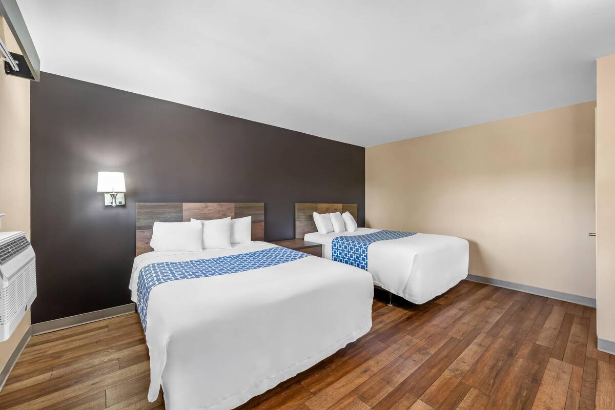 Quarto de hotel com duas camas, uma luminária fixada na parede e piso de madeira.