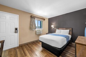 A hotel room with a bed, a desk, a window with curtains, an air conditioning unit, and a door. - Suburban Studios Charlotte - Ballantyne (Charlotte)