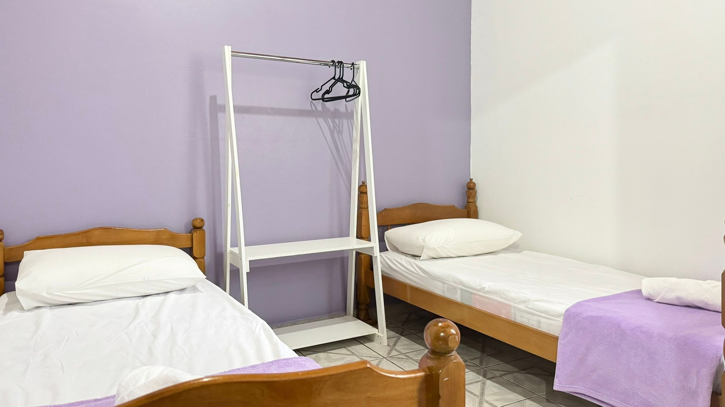 Une pièce avec deux lits en bois, chacun étant équipé de draps blancs et d’une serviette violette. Une étagère métallique blanche est fixée au mur entre les deux lits.