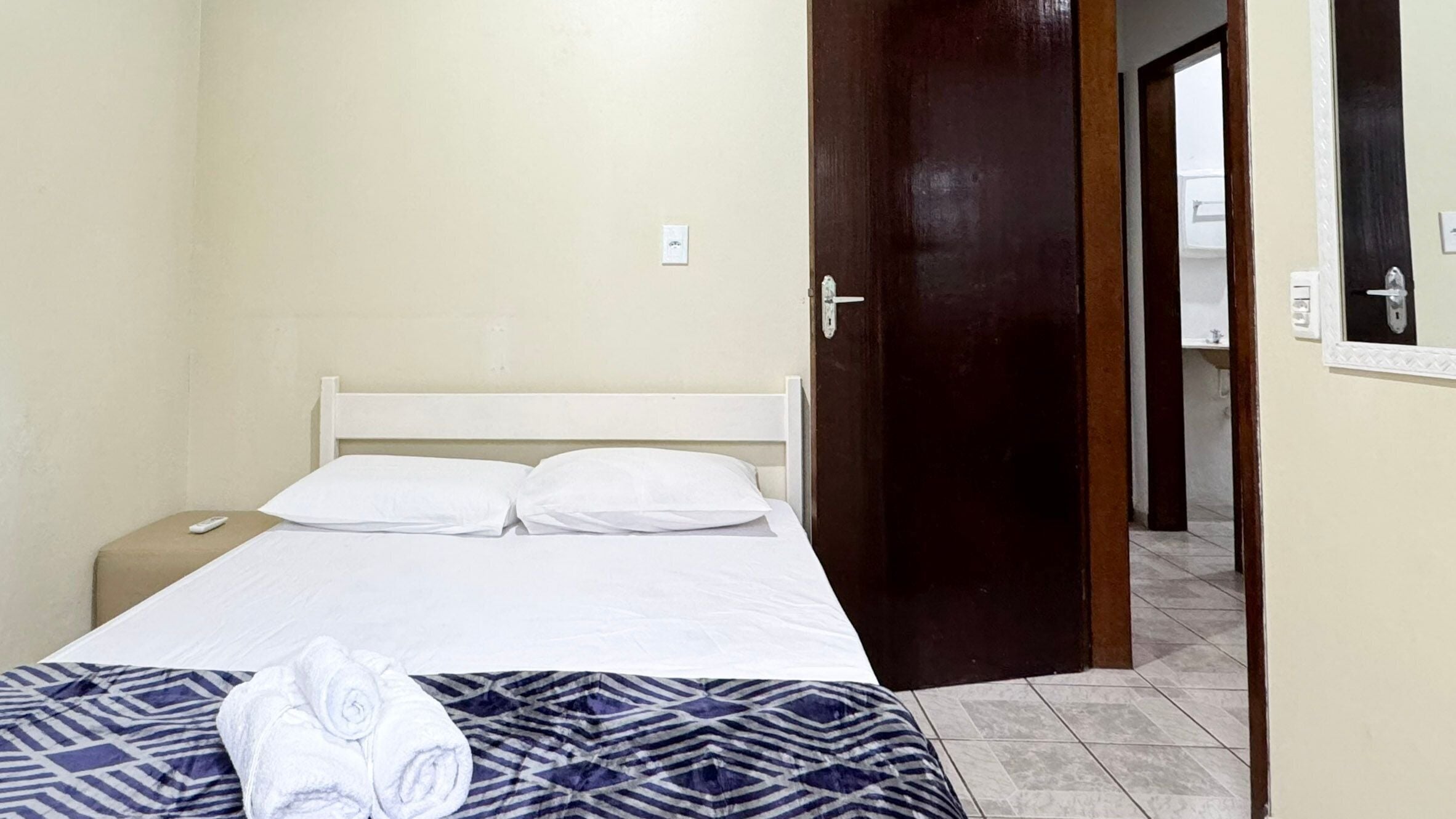 A single bed with white linens, a patterned blanket, and rolled towels on the footboard. A wooden door leads to a bathroom with a mirror and a sink.
