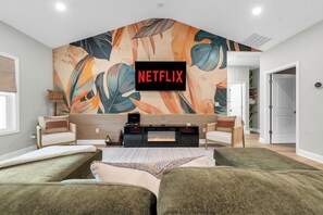 A modern bedroom with a large bed, a fireplace, a wall-mounted TV, and a tropical leaf mural. - Family Retreat, Fire Pit, Games, Close to Ft Moore (Columbus)