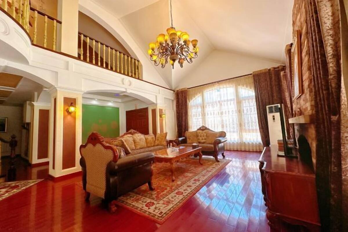Ein großzügiges Wohnzimmer mit hoher Decke, einer Chandelier, einer Sofagarnitur, einem Couchtisch und einem großen Fenster mit Vorhängen.