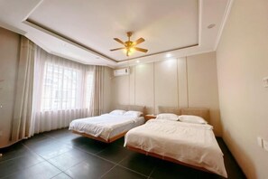 A hotel room with two single beds, a ceiling fan, an air conditioning unit, and a large window with curtains.