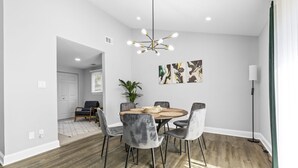 A dining area with a round table, six chairs, a chandelier, and artwork on the wall.